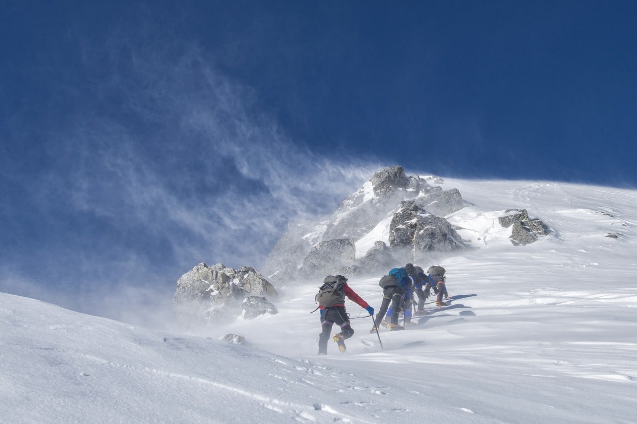 A group of climbers in full expedition gear ascending a steep, snowy mountain slope, pushing forward in challenging conditions.