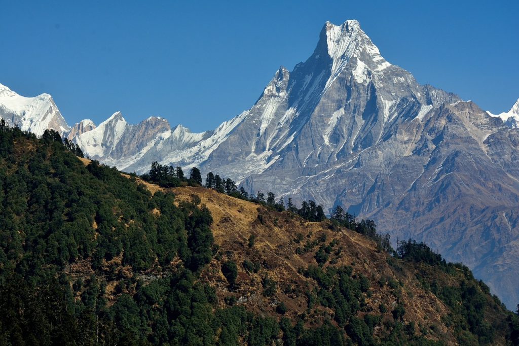 A striking picture of Mount Machhapuchhre, also called the "Fishtail Mountain," with its recognizable snow-covered double peak and a clear blue sky in the background. The rough terrain and smaller mountains in the foreground add to the famed peak's formidable grandeur.