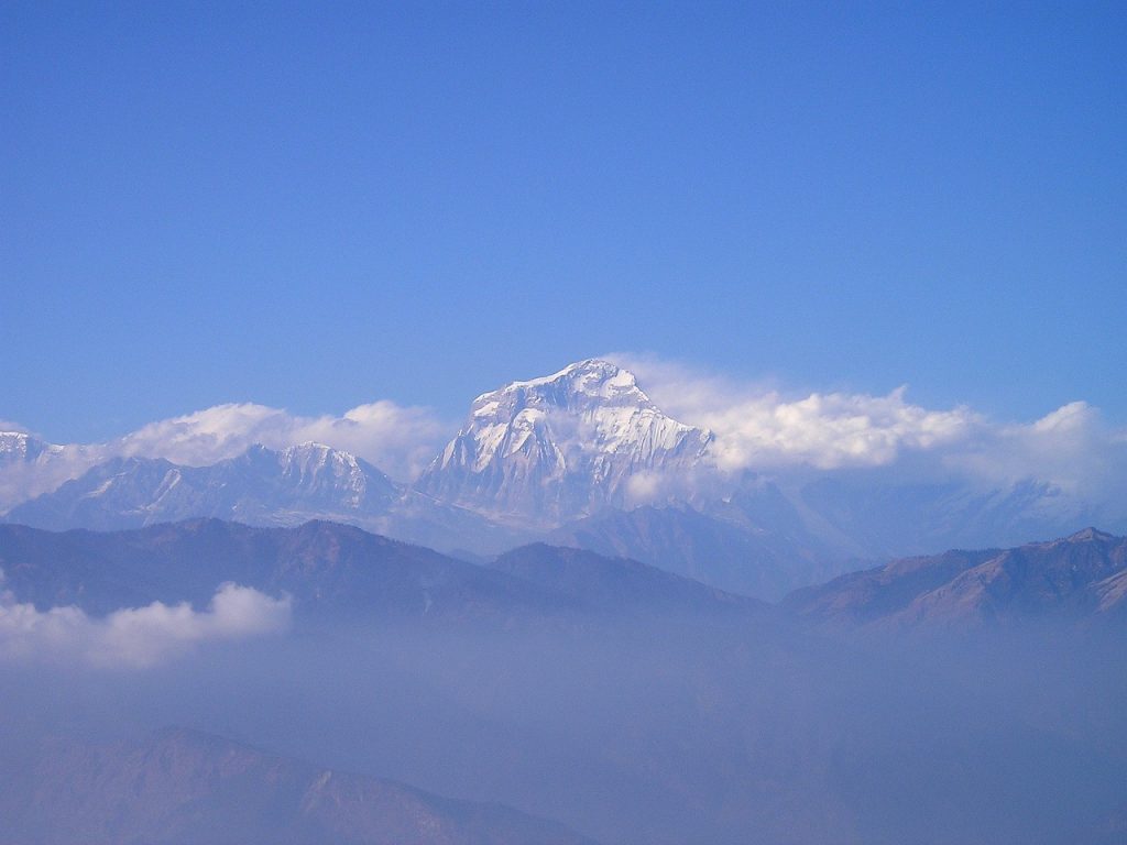 A striking picture of Mount Dhaulagiri, the seventh-highest mountain in the world, with a bright, clear sky in the background and its snow-covered top in the foreground. The craggy slopes and lower hill ranges can be seen in the foreground, highlighting the mountain's stunning contrast and enormous magnitude.