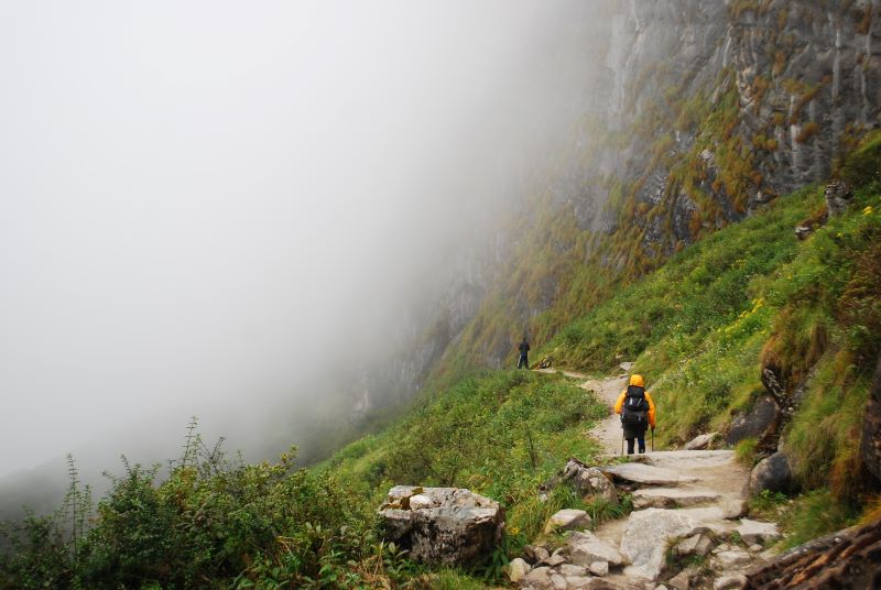 A winding trekking path in the Annapurna region, with distant snow-capped peaks and a clear blue sky overhead.