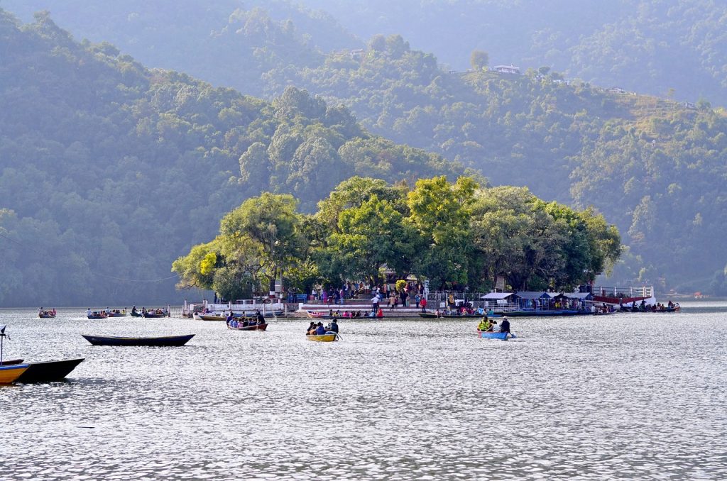 Scenic view of Phewa Lake with calm reflective waters, nestled against forested hills and a distant mountain backdrop.
