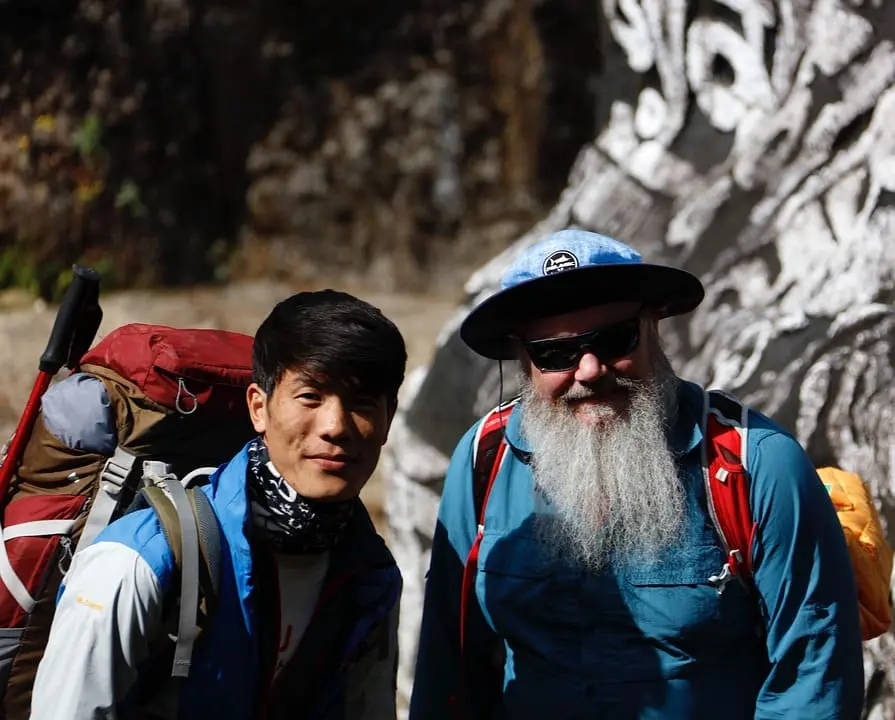 A guide takes a photo of a trekker on a scenic mountain trail, surrounded by Himalayan peaks and clear skies, capturing the essence of the trekking adventure.
