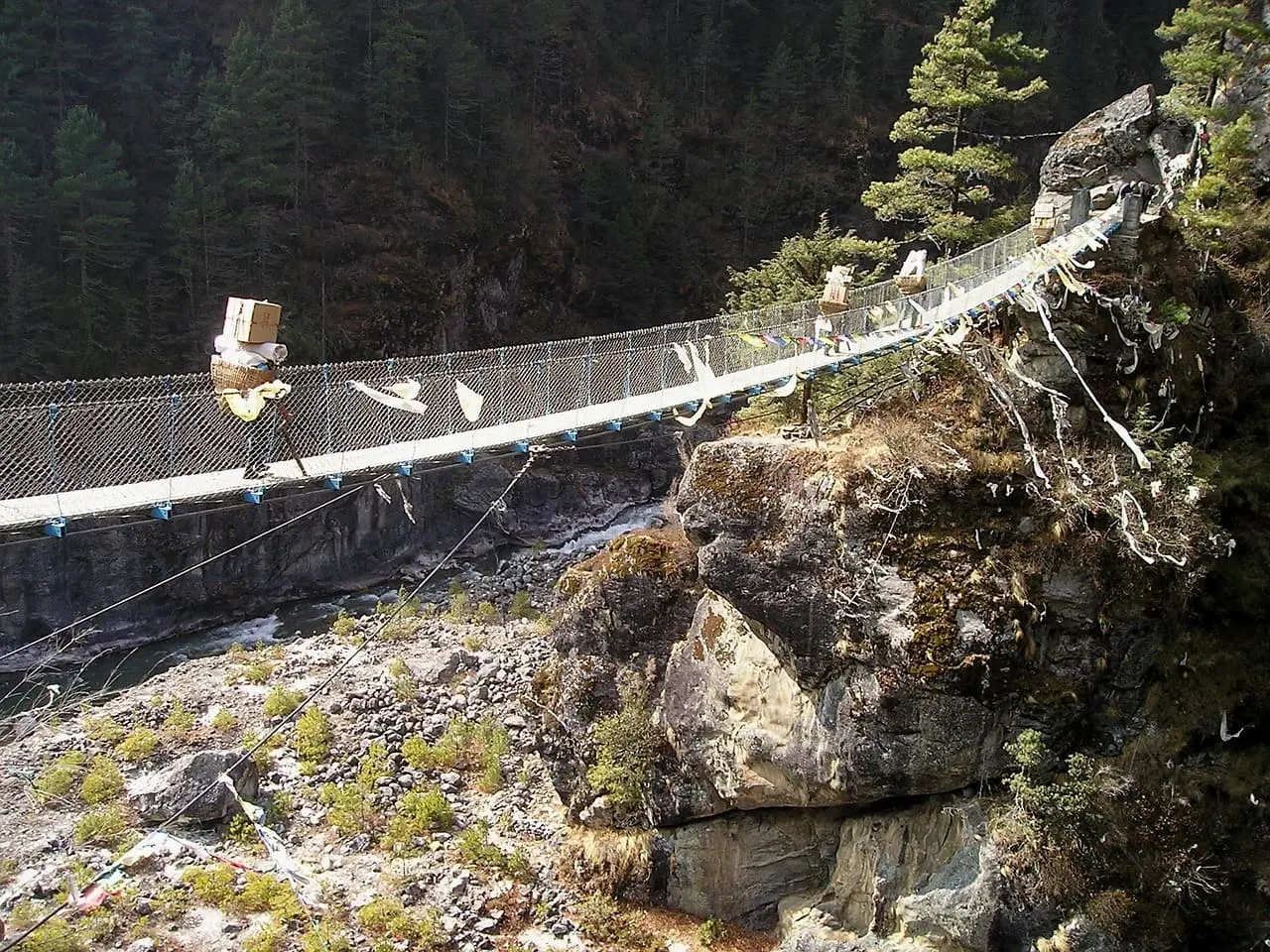 Porters carrying heavy loads carefully walk across a swinging bridge over a river in the Himalayas, showcasing the strength and dedication of local trekking support.